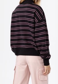 Suéter negro de gran tamaño con rayas horizontales rosas, puños acanalados y cuello alto falso, combinado con pantalones cargo rosa claro.