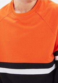 Felpa arancione con collo rotondo, caratterizzata da strisce orizzontali nere e bianche sul fondo. Materiale in cotone morbido e maniche raglan.
