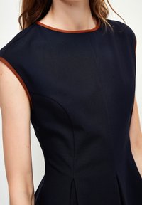 Hihaton tummansininen mekko ruskeilla kursiivireunuksilla kaula-aukossa ja käsiaukoissa, päällä vaaleaihoinen henkilö, jolla on laineikkaat ruskeat hiukset.