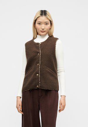 Jeune femme portant un gilet polaire marron boutonné par-dessus un haut blanc à manches longues et un pantalon marron, debout devant un fond uni.