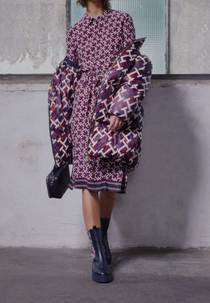 Mujer con vestido y chaqueta acolchada con patrón a juego en rojo, blanco y azul, llevando un pequeño bolso negro y botas oscuras.