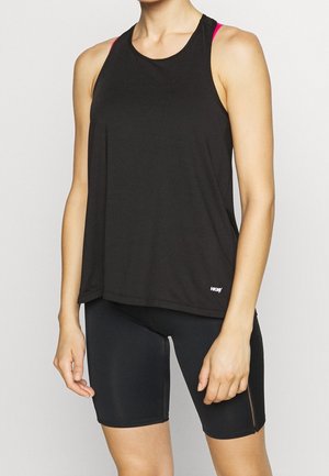 Frau trägt ein schwarzes, ärmelloses, locker sitzendes Top über einem rosa Sport-BH und schwarze, enganliegende Radlerhosen, steht vor einem einfarbigen Hintergrund.