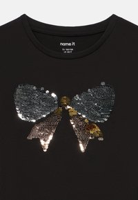 T-shirt preta com um grande design de laço feito de lantejoulas em prata e ouro. Decote redondo e textura de tecido suave. Tamanho EU 122/128.