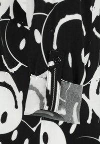 Black and white fabric featuring large circular patterns with a textured finish, includes a button and zipper detail on the cuff.