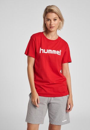 Red cotton t-shirt featuring a prominent white "hummel" logo, paired with grey shorts. The outfit has a casual, comfortable design.