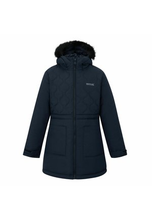 Lächelndes Mädchen mit langen blonden Haaren trägt eine dunkelblaue Parka, weißes Poloshirt, schwarzen Rock und hält eine blaue Wasserflasche, stehend vor einfachem Hintergrund.