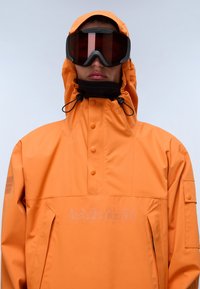 Person med en orange hættetrøje med frontknapper og sorte skibriller foran en simpel lys baggrund.