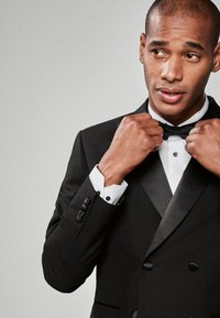 Sort smoking med satin revers, kombineret med en hvid skjorte og sort butterfly. Modellen justerer butterflyen og fremviser knappedetaljerne.