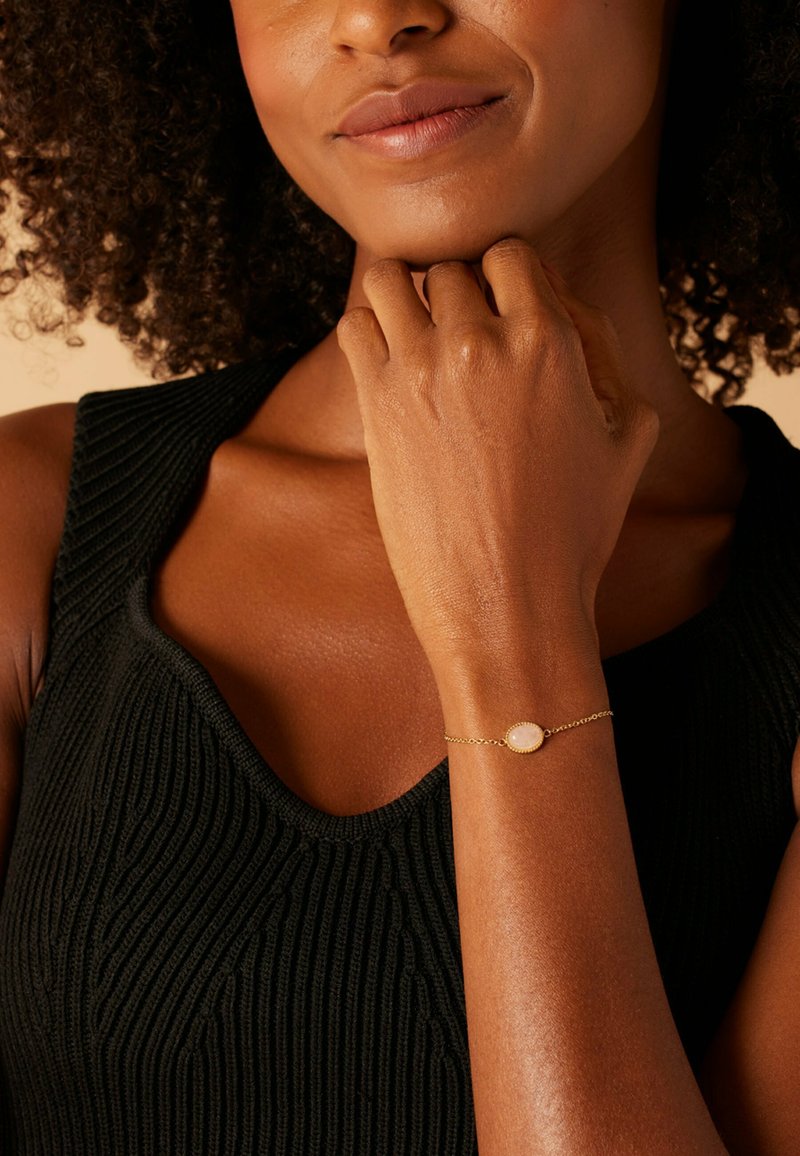 Gouden kettingarmband met een ovale, doorschijnende steeninstelling. Het model draagt een getextureerde zwarte tanktop, waardoor de armband op haar pols goed zichtbaar is.