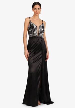 Frau in einem schwarzen Satin-Abendkleid mit tiefem V-Ausschnitt, verziert mit silbernen Pailletten, trägt Perlenohrringe und schwarze Sandalen.