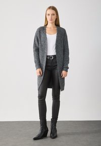 Grijze gebreide cardigan met een open voorkant, lange mouwen en geribbelde manchetten, gedragen over een wit shirt en gecombineerd met zwarte leren broeken en laarzen.