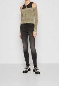 Mujer con un top verde salvia sin hombros con cremallera, una camiseta negra debajo, jeans ajustados negros desgastados y zapatos de plataforma plateados con suelas negras.