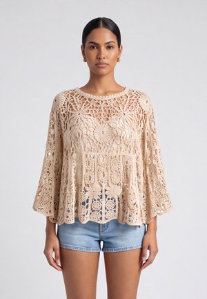 Vrouw die een beige gehaakte kanten top met bloemenpatronen draagt en blauwe denim shorts, staand tegen een effen lichte achtergrond.