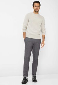 Hellgrauer gestrickter Pullover mit gerippter Textur, gepaart mit dunkelgrauen maßgeschneiderten Hosen und schwarzen Lederschuhen. Das Modell steht mit den Händen in den Taschen.