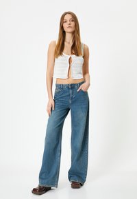 Biały, dziergany crop top z dekoltem w kształcie klucza; dżinsy niebieskie o wysokim stanie i szerokich nogawkach; model noszący brązowe buty na platformie.