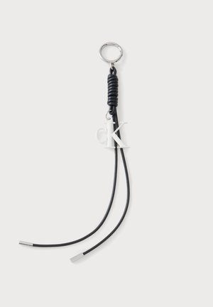Sleutelhanger met zwart opgerold koord, twee losse uiteinden met zilveren uiteinden en een wit "cK" logo bedeltje dat onder de metalen ring hangt.