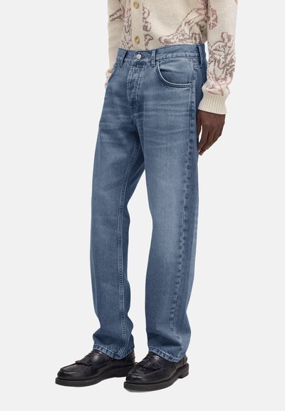 Personne portant un jean droit bleu clair, des mocassins en cuir noir et un pull en tricot beige avec un motif.