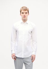 Chemise blanche avec une patte de boutonnage comportant des plis verticaux, col classique et manches longues. Portée avec un pantalon gris clair.