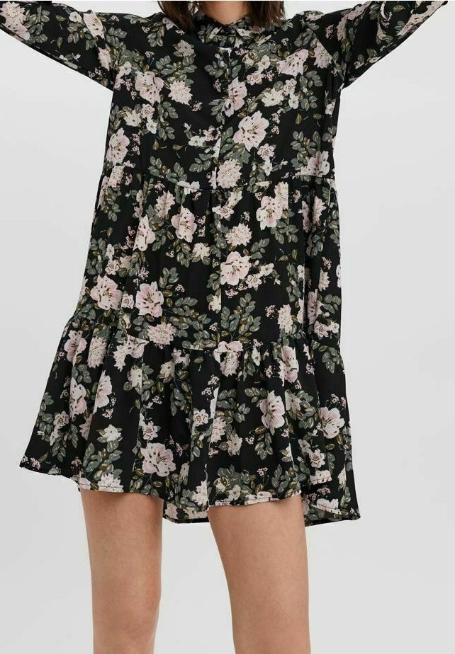 Femme portant une robe noire à fleurs à manches longues avec des motifs floraux roses et verts et un ourlet à volants, les bras levés au-dessus de sa tête.