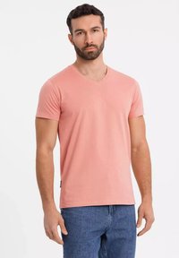 T-shirt a scollo a V di un delicato rosa chiaro, realizzata in cotone, con maniche corte, design semplice, indossata con jeans blu, presenta un piccolo etichetta sul lato.