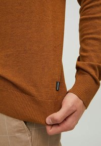 Brun stickad tröja med ribbad nederdel och ärmslut, med en liten svart etikett på sidan. Närbild av en hand som håller i nederdelen.