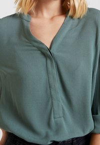 Frau trägt eine lockere, langärmelige grüne Bluse mit einem V-Ausschnitt, die in dunkle hochgeschnittene Hosen gesteckt ist, vor einem neutralen Hintergrund.