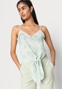 Camisola em tie-dye verde-claro com alças finas e decote em V, apresentando um detalhe frontal amarrado. Combinada com calças leves verdes-claro.