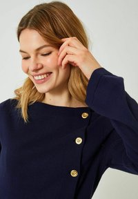 Marinblå topp med långa ärmar, som har en diagonal knapplist med fyra gyllene knappar och en rund halsringning. Mjuk tygkvalitet.