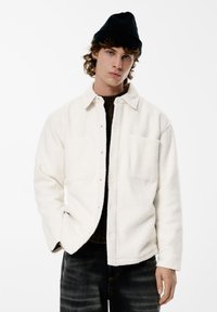 Valkoinen fleece-takki, jossa on napillinen etuosa, kaksi rintataskua ja kaareva helma, pidetty tumman neuleen päällä ja yhdistetty tummiin farkkuihin. Musta pipo.