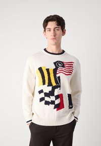 Pull en tricot crème avec des motifs de drapeau multicolore et à carreaux, col et poignets côtelés, avec un petit logo en bas.