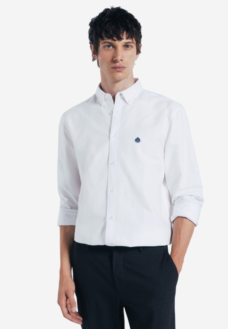 Camicia bianca con bottoni, dotata di un piccolo logo blu sul lato sinistro del petto, con maniche lunghe arrotolate fino ai gomiti e una texture in cotone liscia.