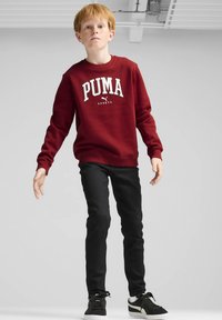 Sudadera color burdeos con el logo "PUMA SPORTS" en blanco, combinada con pantalones negros de corte slim y zapatillas negras con suelas blancas y detalles verdes.