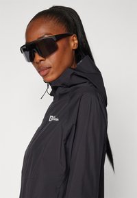 Black waterproof jacket with hood and logo on chest; worn with oversized black sunglasses, highlighting a sleek design and modern aesthetic.