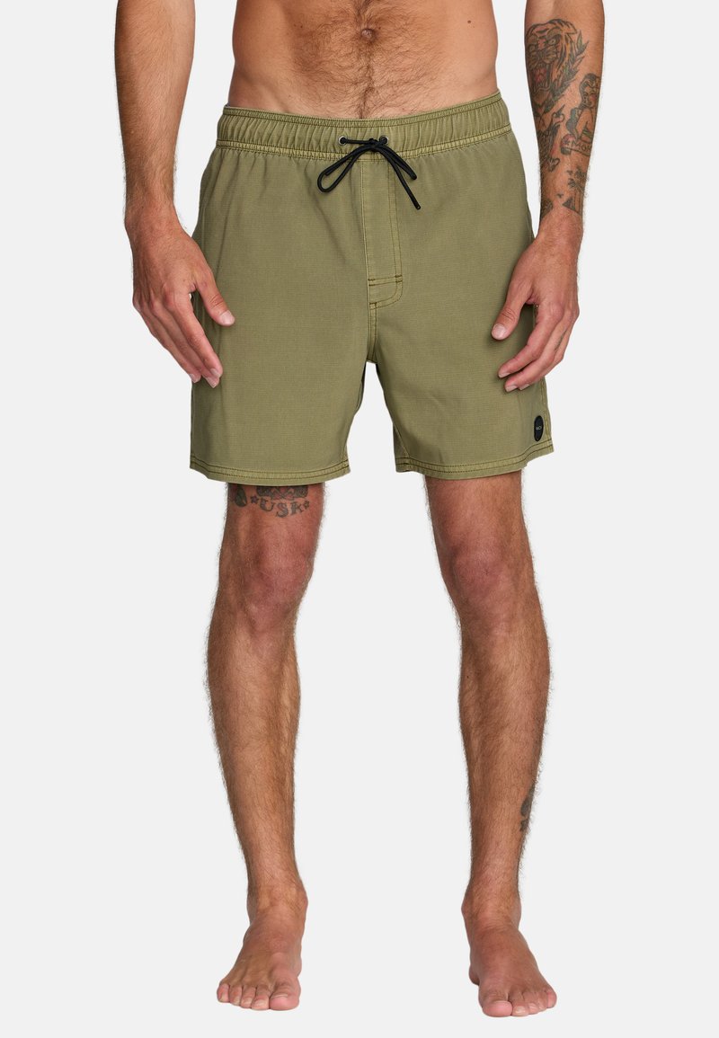 Mann trägt olivgrüne Badeshorts mit schwarzer Kordel, steht barfuß und zeigt sichtbare Arm- und Beintattoos, dargestellt vom Taillenbereich bis zu den Füßen.