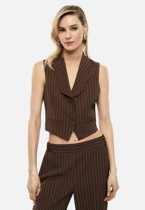 Donna che indossa un gilet blazer senza maniche marrone a righe sottili abbinato a pantaloni a vita alta dello stesso tessuto, su uno sfondo uniforme.