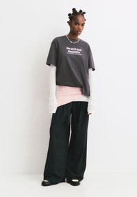 Jongere met gestyled haar, gekleed in een gelaagde oversized grijze T-shirt met tekst, witte lange mouwen, een roze omslag, brede zwarte broek en chunky schoenen.