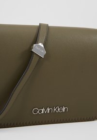 Clutch in pelle verde oliva con logo silver di Calvin Klein, caratterizzato da un design elegante, una texture liscia e dettagli di cucitura a contrasto.