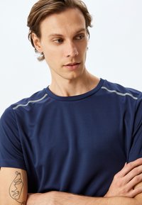 Mörkblå atletisk t-shirt gjord av texturerat tyg, med kort ärm och reflekterande grå detaljer över axlarna.