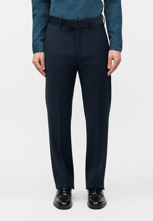 Uomo con pantaloni eleganti blu scuro, scarpe eleganti nere e camicia teal con bottoni e maniche abbassate, in piedi davanti a uno sfondo semplice.