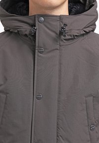 Giacca isolante grigio scuro con colletto alto, chiusura con zip, bottoni a scatto e tasche laterali. Realizzata in materiale testurizzato e resistente alle intemperie.