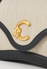 Sac en toile beige avec des bordures en cuir noir, présentant un fermoir central en forme de C de couleur dorée et des détails de coutures ondulées.