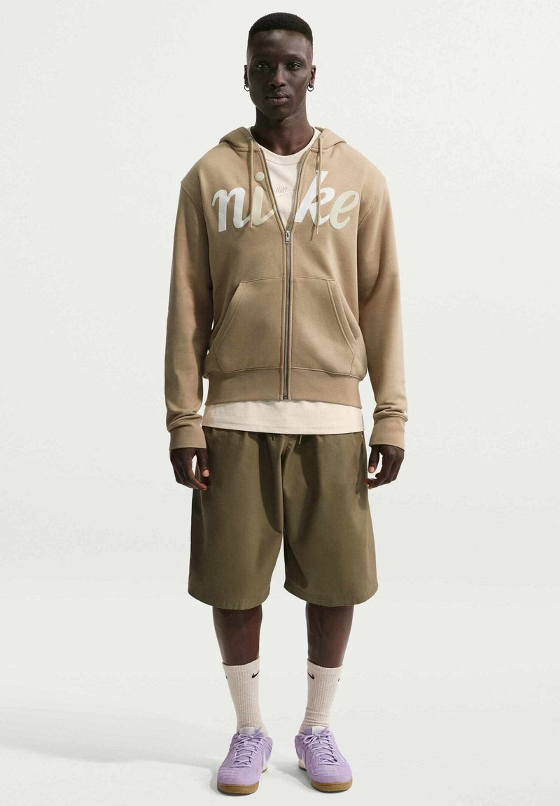 Man stands facing forward wearing a tan Nike hoodie, olive shorts, white socks with a Nike logo, and lavender sneakers on a plain background.