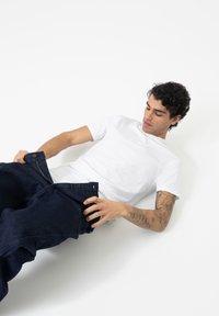 T-shirt blanc �à manches courtes associé à un jean en denim bleu foncé, ample, avec fermeture éclair et bouton, porté de manière décontractée.