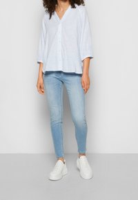 Jasnoniebieska koszula w paski z guzikami, dekoltem w serek i rękawami do łokci, zestawiona z jasnoniebieskimi dopasowanymi dżinsami i białymi trampkami.