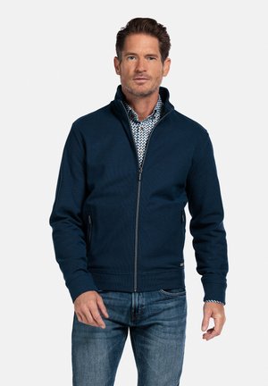 Marineblauwe ritsjack met hoge kraag, gestructureerde stof en zijzakken, gecombineerd met een patroonshirt en blauwe spijkerbroek.