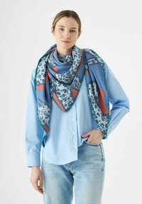 Écharpe multicolore présentant une base bleue avec des motifs rouges, gris et noirs. Bordée de couture verte, drapée sur une chemise bleu clair.