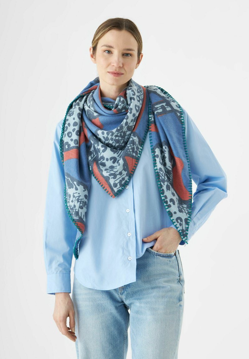 Écharpe multicolore présentant une base bleue avec des motifs rouges, gris et noirs. Bordée de couture verte, drapée sur une chemise bleu clair.