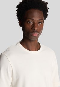 Ung sort mand med kort krøllet hår iført en enkel hvid t-shirt med rund hals, som kigger lige frem med en neutral mine.