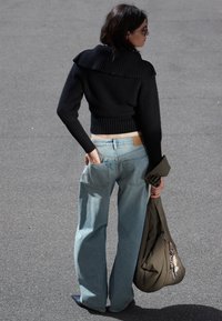 Schwarzer gerippter, verkürzter Pullover mit großem Kragen, hellblaue weite Jeans, schwarze spitze Schuhe und eine olivgrüne Tasche.