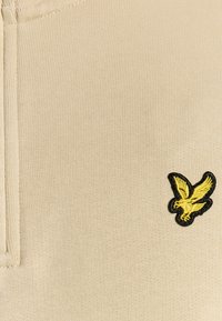 Beige Strickpolo mit Reißverschluss am Kragen und einem gelben, gestickten Vogel-Logo auf der linken Seite. Glatte Textur, figurbetontes Design.